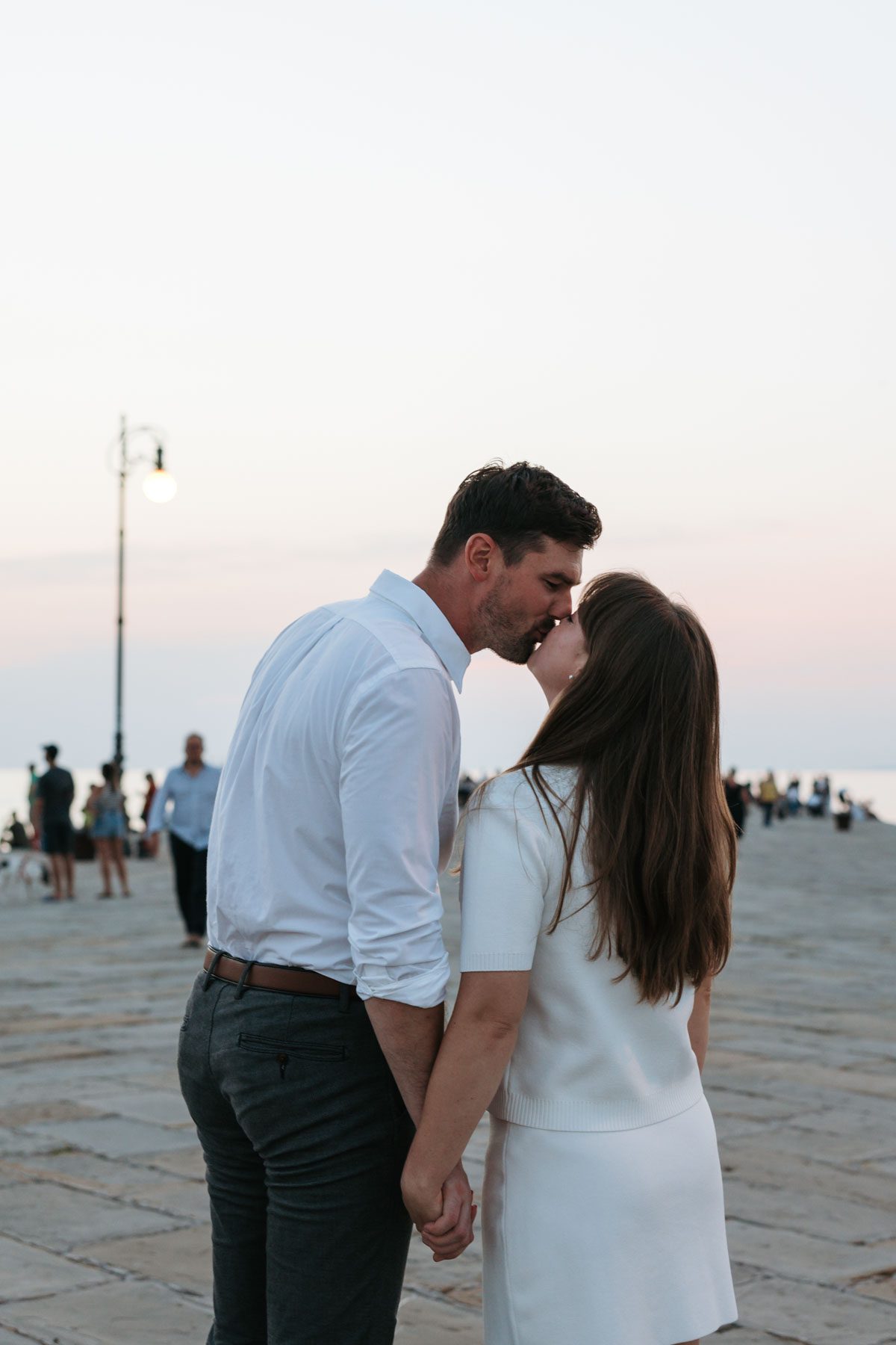 Couple kissing and holding hands during a romantic sunset walk in Trieste, Italy with soft evening light and people in the background