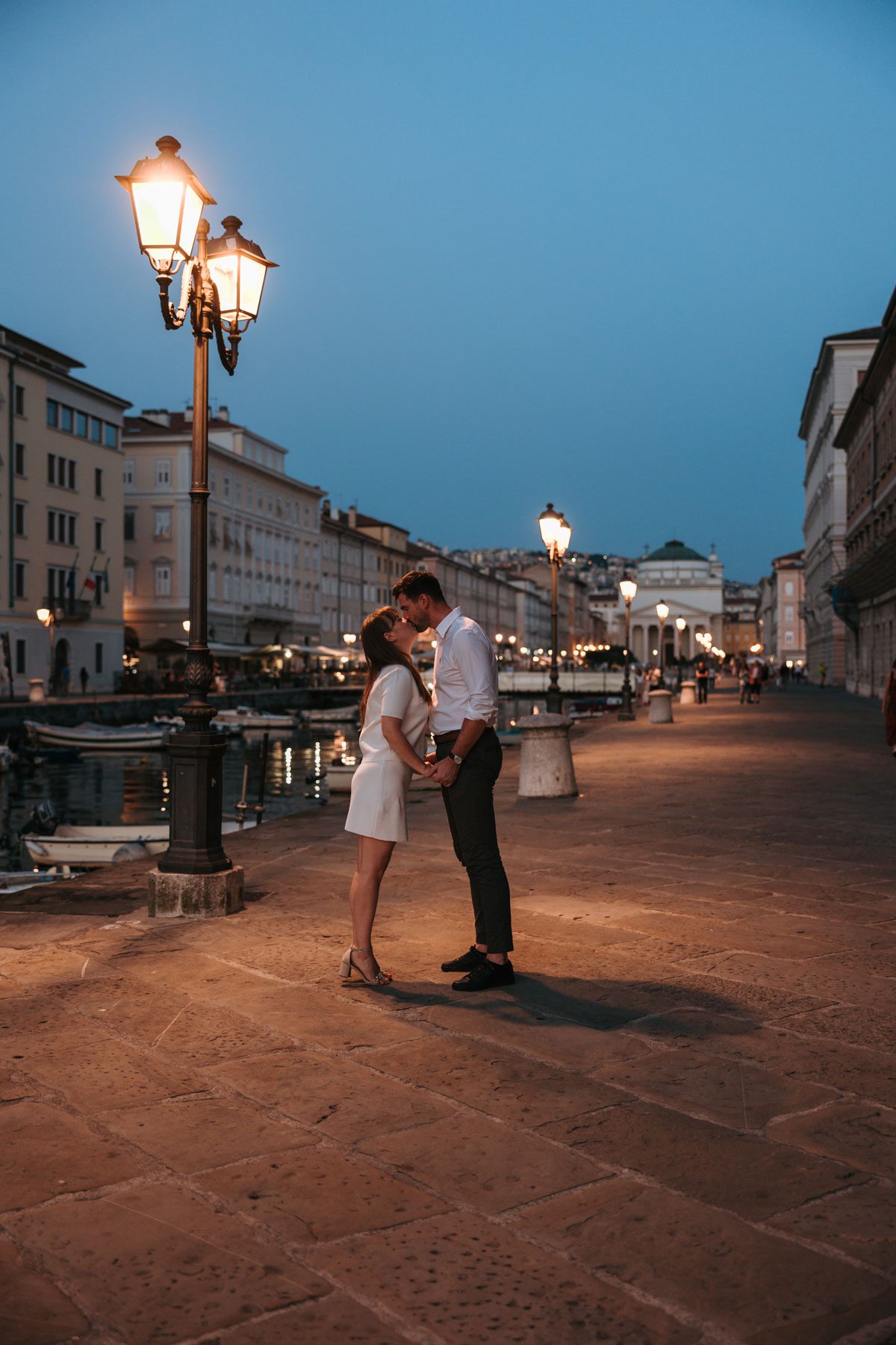 Couple standing close and holding hands under street lights in Trieste, Italy during blue hour with historic waterfront buildings in the background