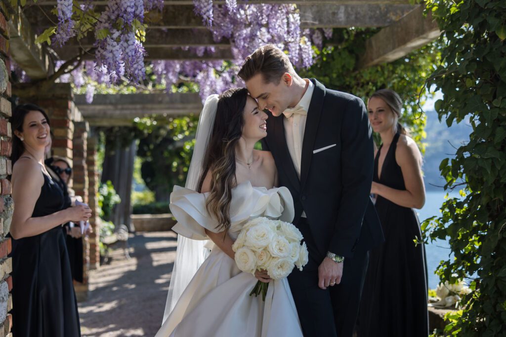 joyful bride and groom walking under wisteria with guests during wedding in Italy
