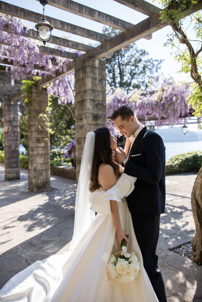 elegant bride with flowing gown and groom under wisteria pergola by the sea in Italy