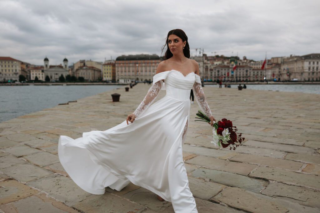 Elegant bride walking by the sea in Trieste, Italy wearing a flowing wedding dress and holding a bouquet during a luxury bridal editorial shoot