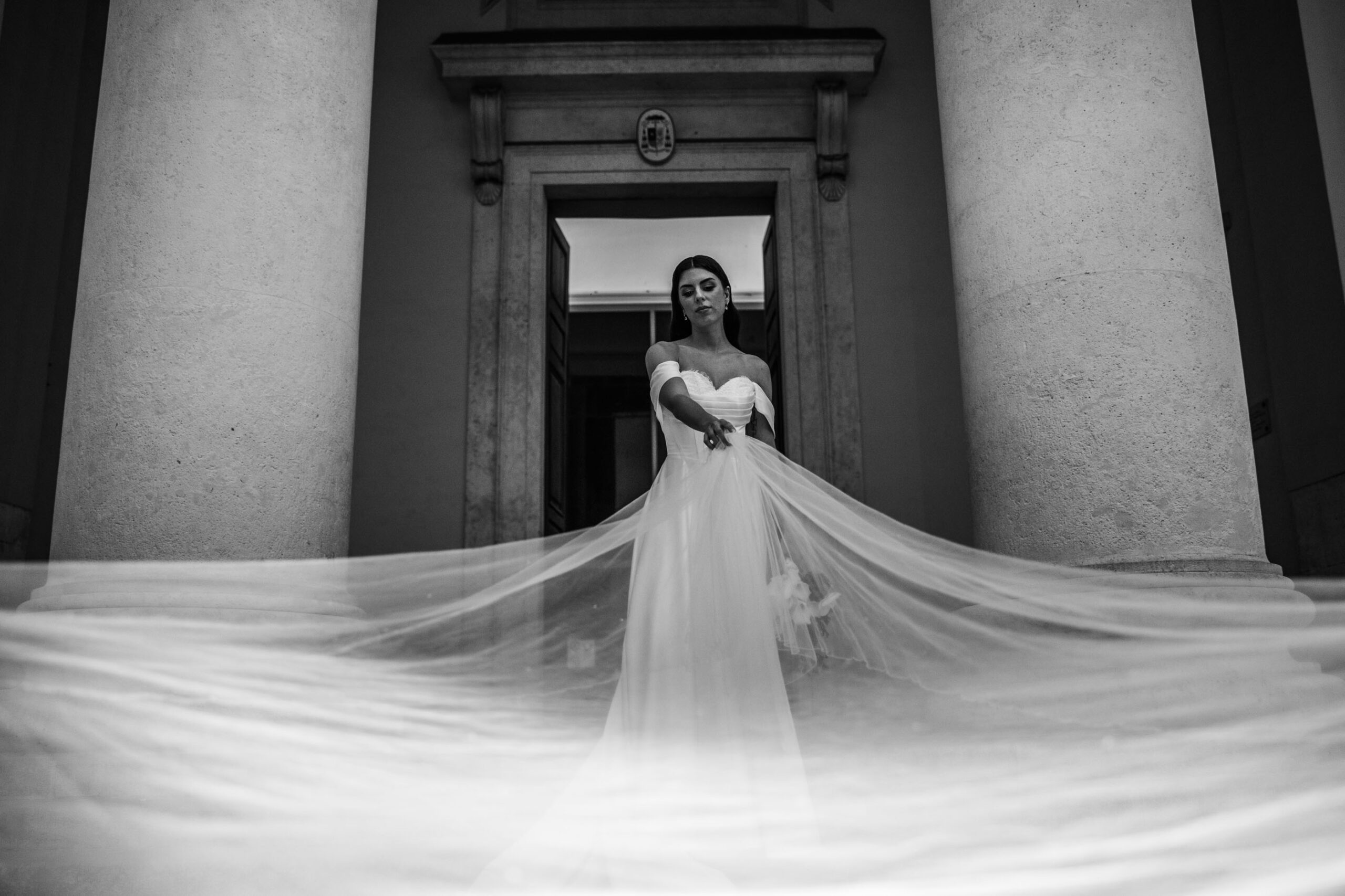 Luxury bridal editorial in Trieste, Italy with a bride and flowing veil in motion in front of neoclassical architecture creating a dramatic and elegant scene