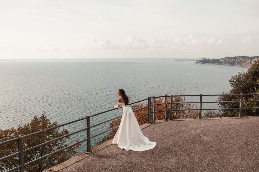 Bride in a flowing wedding dress overlooking the Adriatic coast near Trieste, Italy during a romantic destination bridal session.
