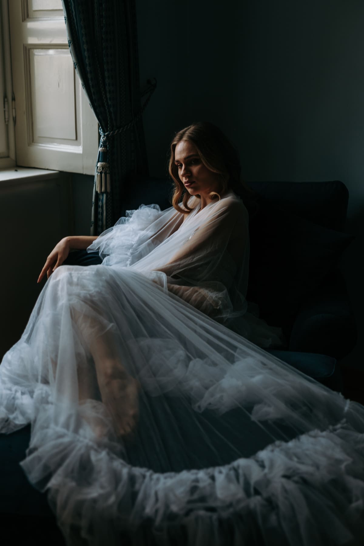 Bride resting on a sofa wrapped in soft tulle, quiet and emotional wedding morning moment with natural light