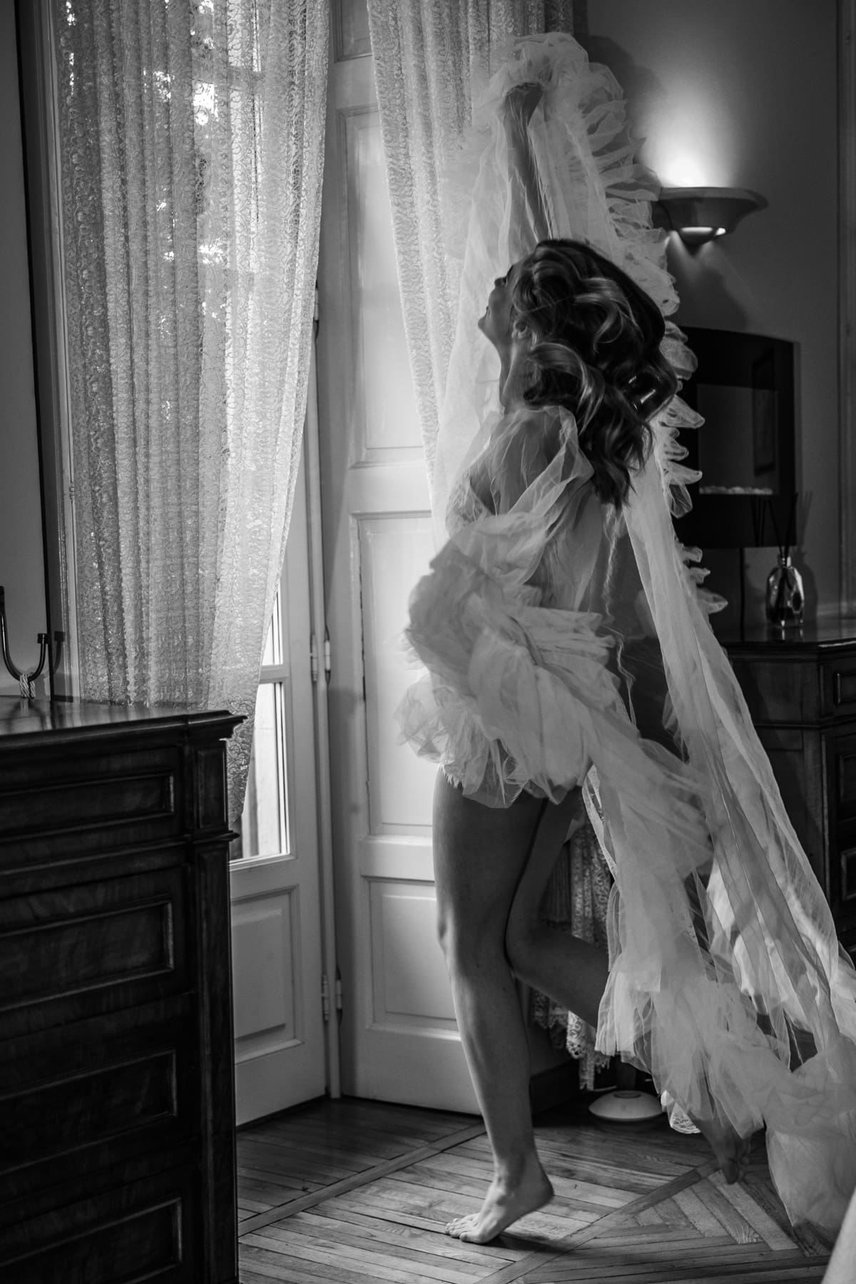 Bride in flowing veil standing by the window, soft natural light creating an intimate wedding morning atmosphere in Italy