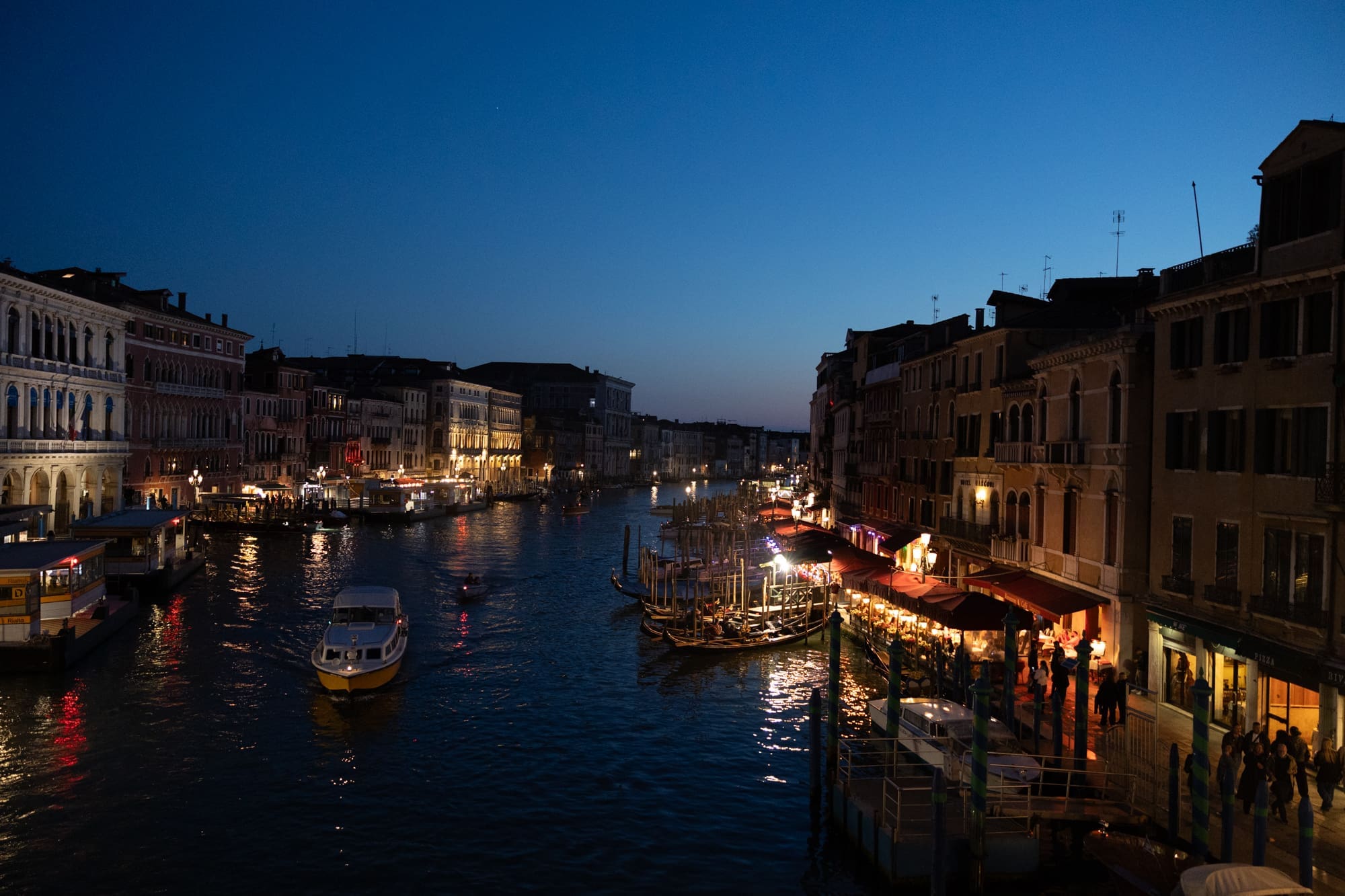 Venice at night along the Grand Canal near Rialto bridge with glowing lights, boats and historic buildings