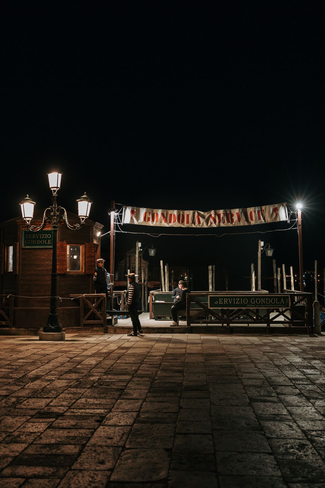 Historic gondola station in Venice at night with glowing lanterns and gondoliers waiting by the canal, cinematic atmosphere near Piazza San Marco in Italy.