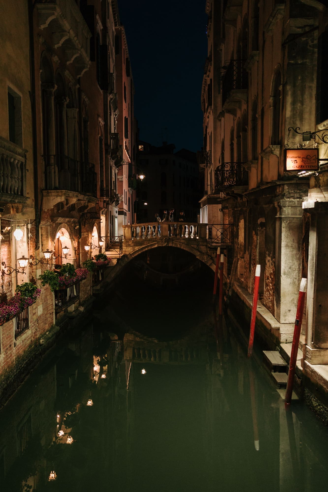 Quiet Venice canal at night with romantic bridge, glowing lanterns, and historic Italian architecture reflected in the water near Piazza San Marco.