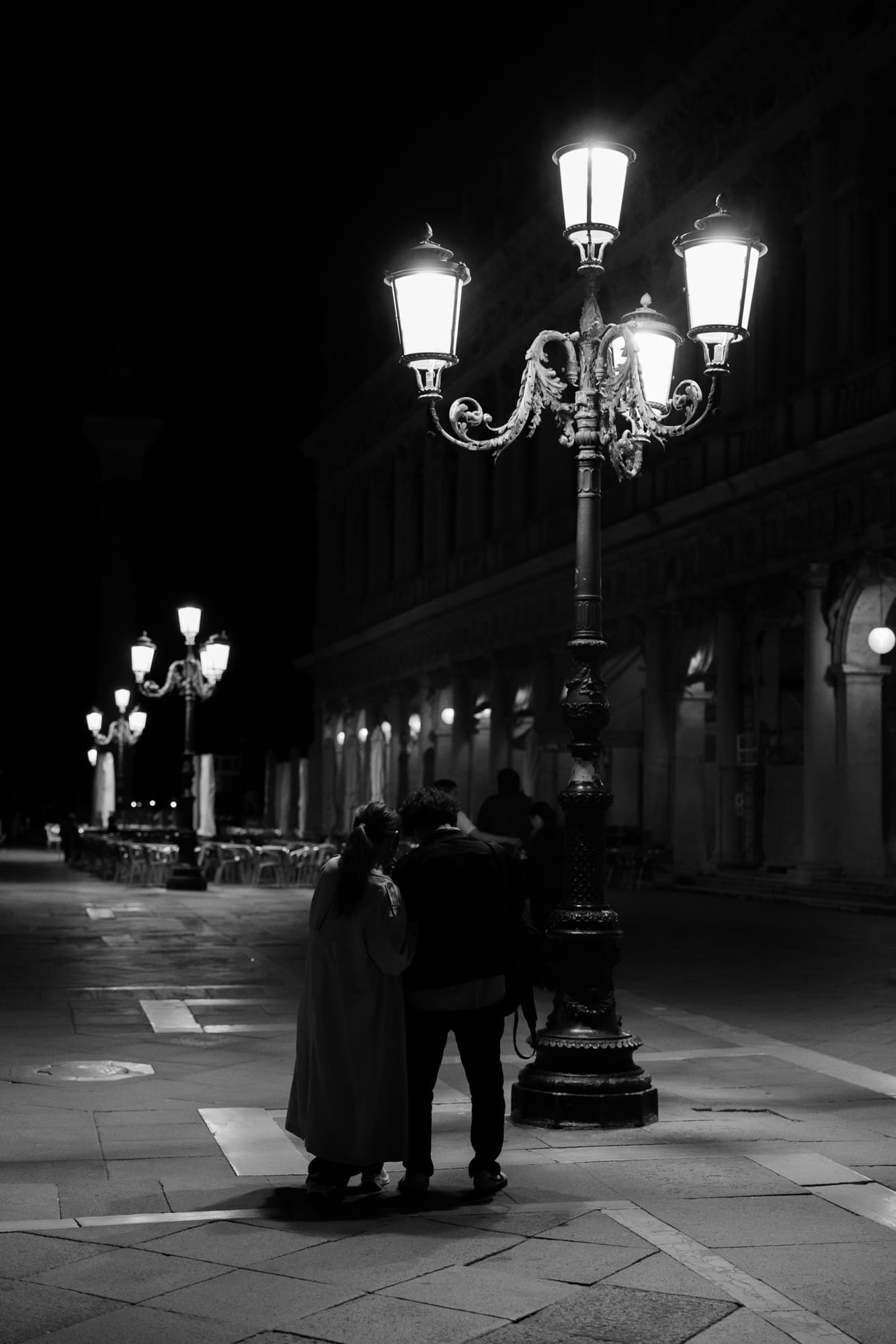 Romantic couple walking through quiet Venice at night beneath glowing historic street lamps near Piazza San Marco, black and white editorial elopement photography in Italy.