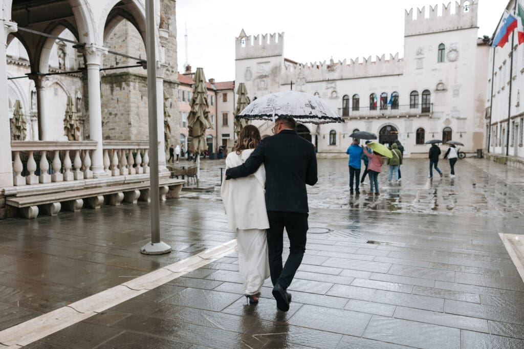 Bride and groom walking across Tito Square in Koper, Slovenia, with the historic Praetorian Palace in the background where they originally planned to hold their civil wedding ceremony.