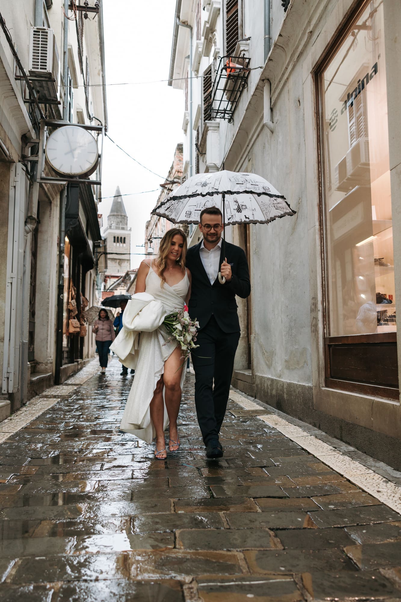 Bride and groom walking along Čevljarska Street in Koper, Slovenia, with the bell tower of the Cathedral of the Assumption visible in the background.