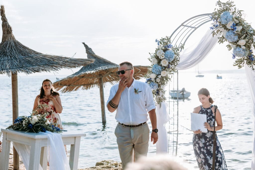 Emotional groom during a beach wedding ceremony on Levan Island in Croatia standing beneath a floral arch by the Adriatic Sea.