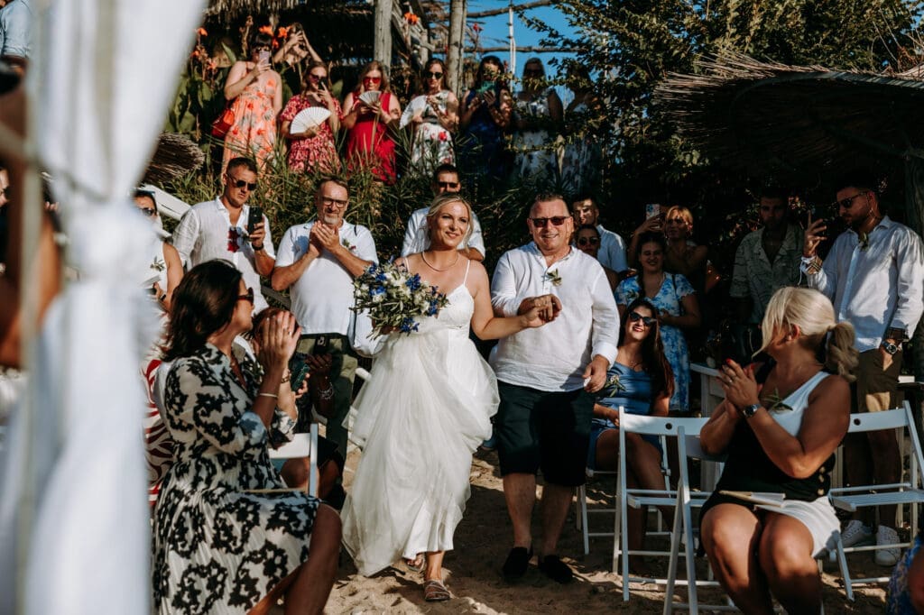 Bride entering her beach wedding ceremony with her father surrounded by guests on Levan Island in Croatia during golden hour.