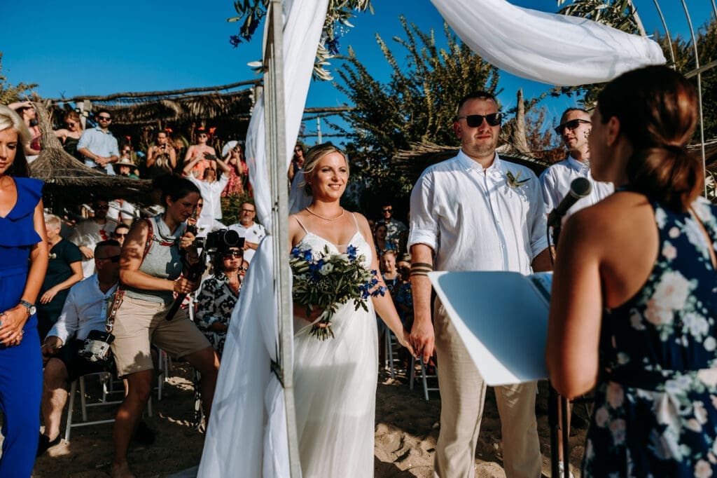 Bride and groom standing beneath a floral arch during an intimate beach wedding ceremony on Levan Island in Croatia surrounded by family and guests.
