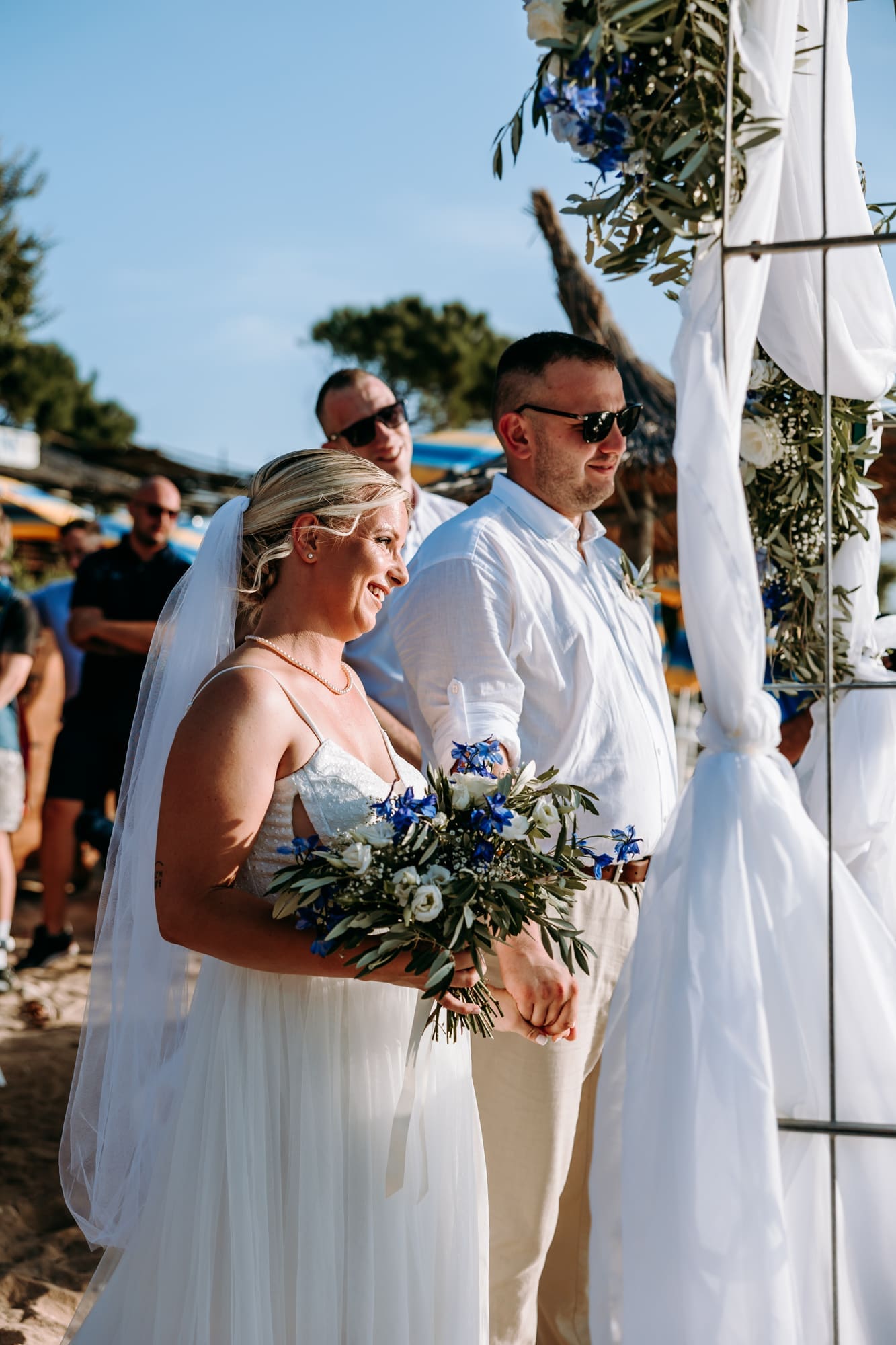Bride and groom smiling during their intimate beach wedding ceremony on Levan Island in Croatia while holding hands beneath a floral arch by the sea.