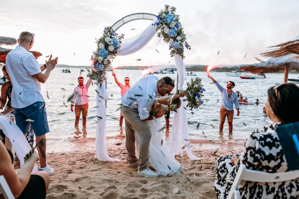 Bride and groom kissing beneath a floral arch during their beach wedding ceremony on Levan Island in Croatia while guests celebrate with smoke flares by the Adriatic Sea.