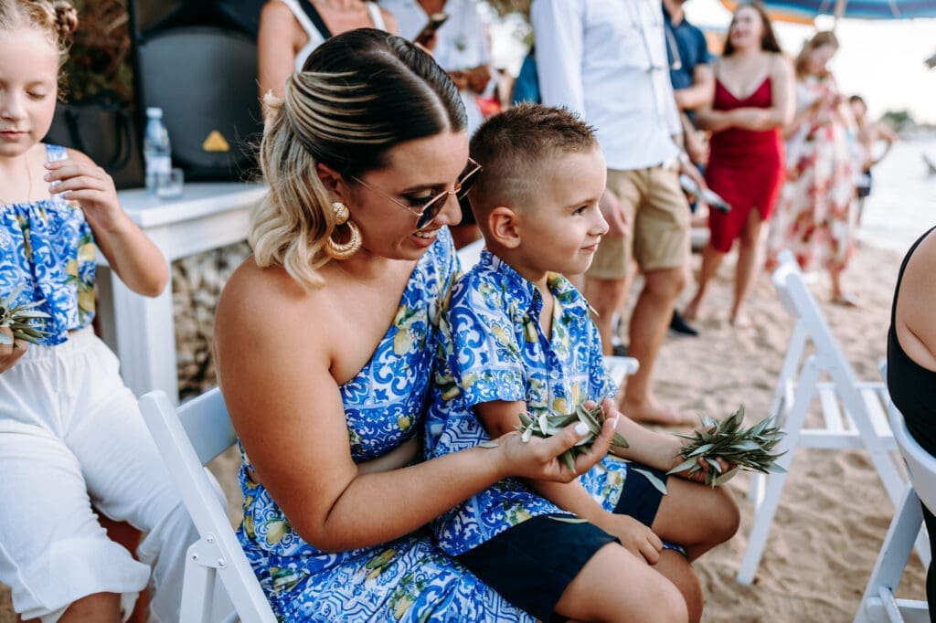 Mother and child sitting together during a relaxed beach wedding celebration on Levan Island in Croatia holding olive branches.