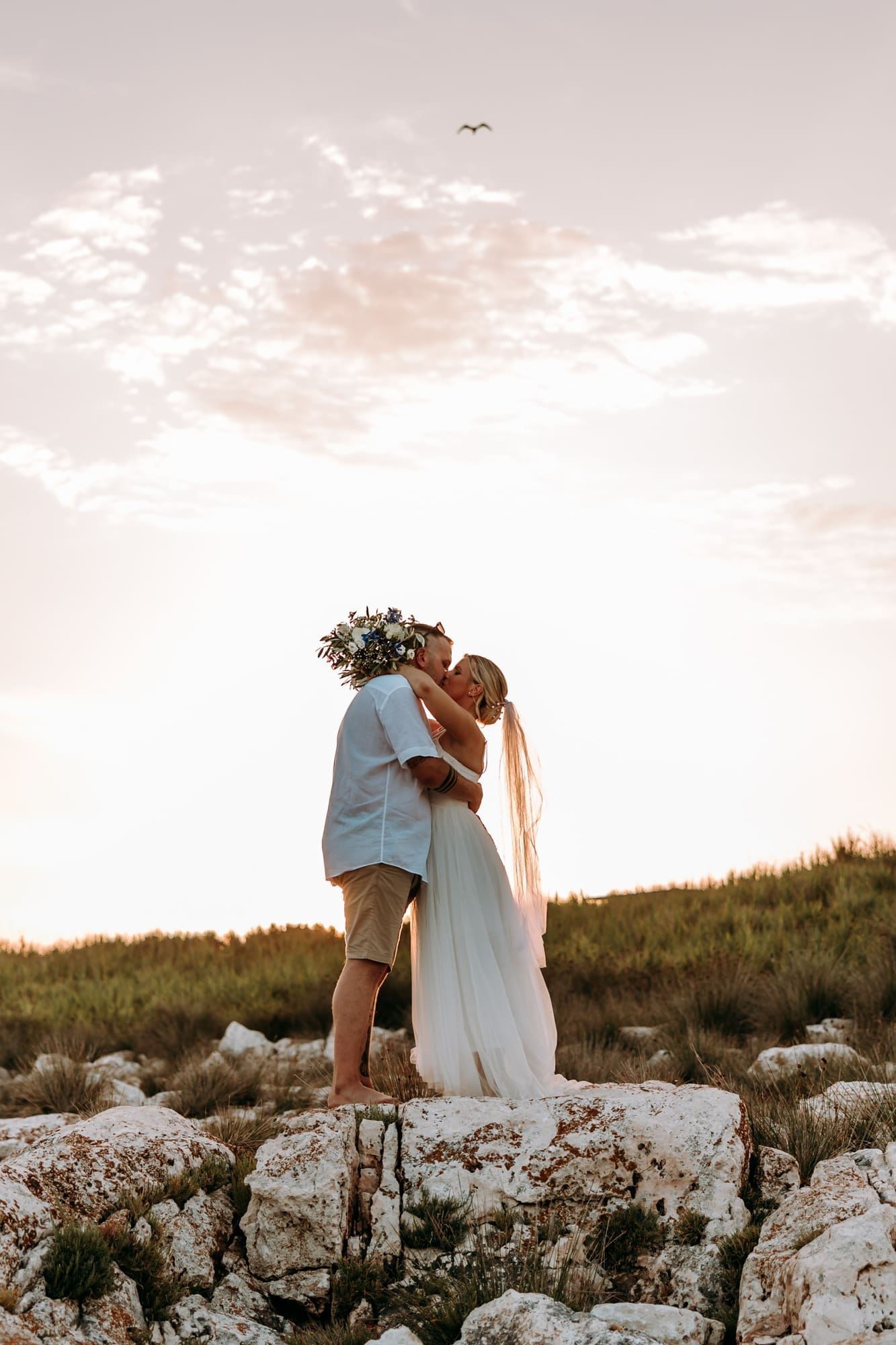 Bride and groom kissing on the rocky coastline of Levan Island in Croatia during a romantic sunset portrait session after their beach wedding ceremony.