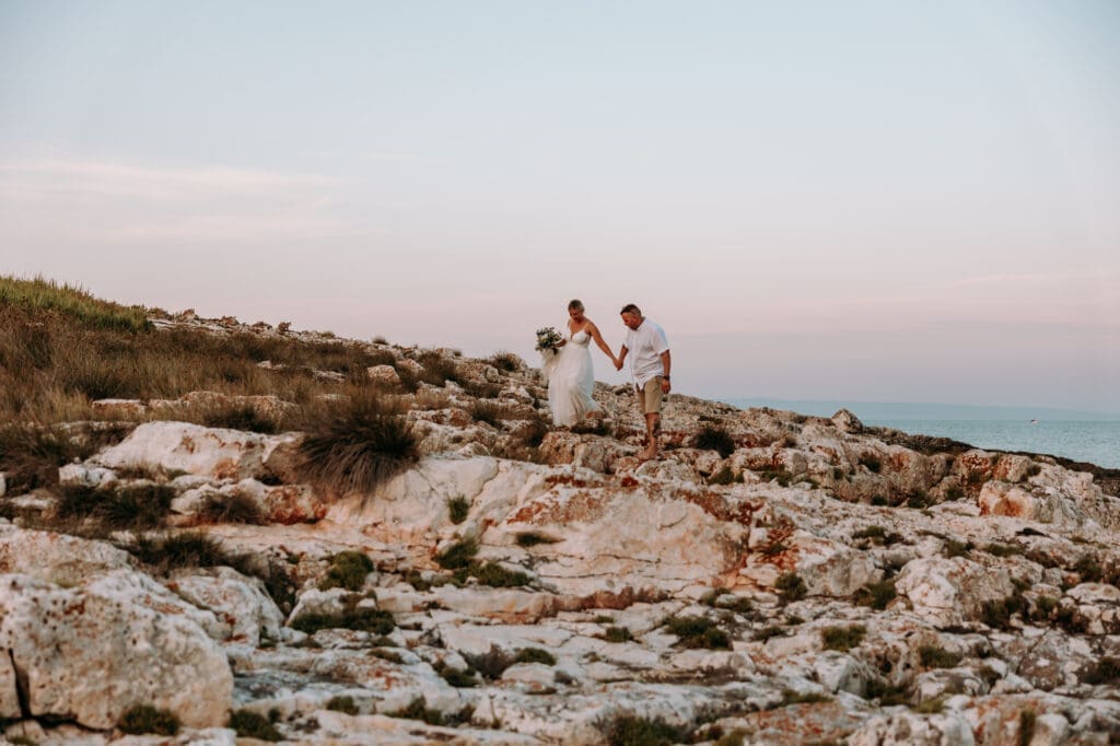 Bride and groom walking across the rocky coastline of Levan Island in Croatia during sunset after their intimate beach wedding ceremony.