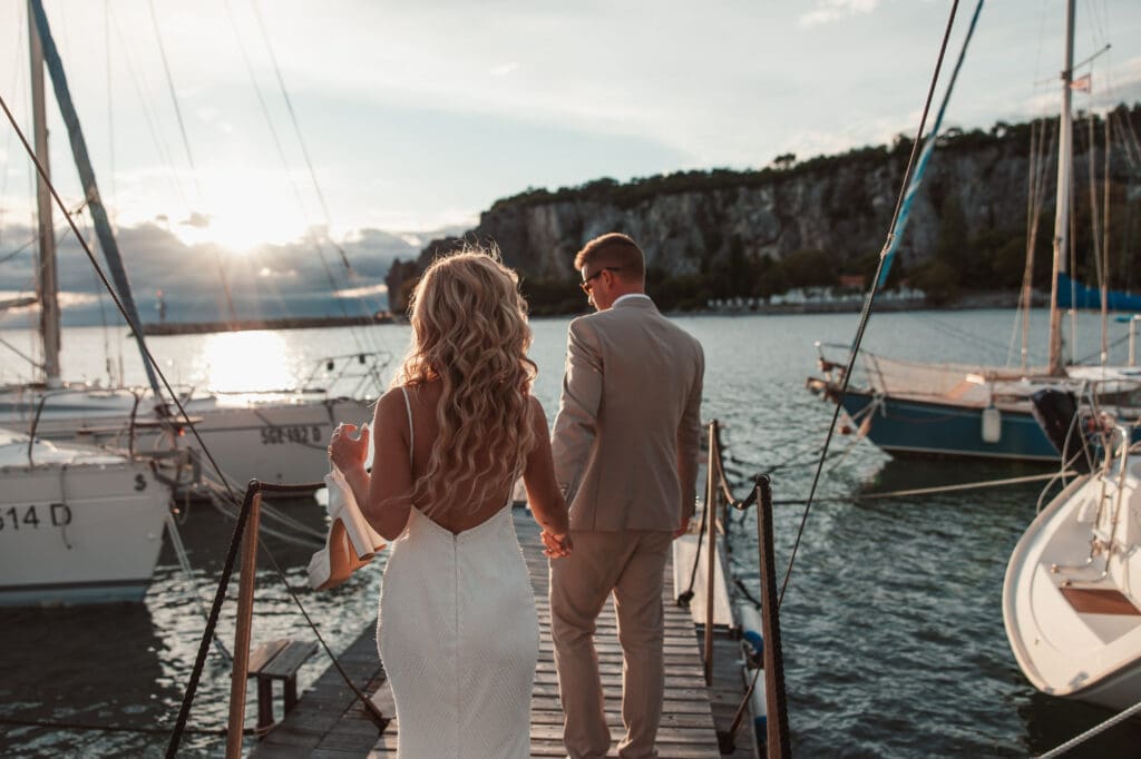 Zala and Matej walking hand in hand along the marina dock during wedding portraits in Sistiana, Italy.