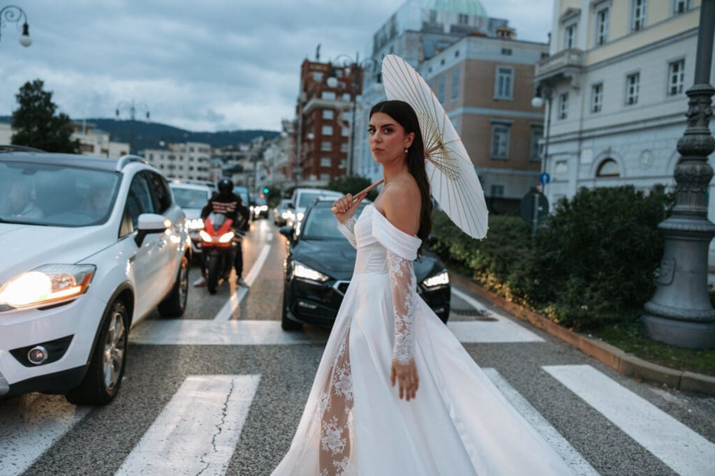 Bride crossing a street in downtown Trieste with a parasol during an editorial wedding photoshoot in Italy