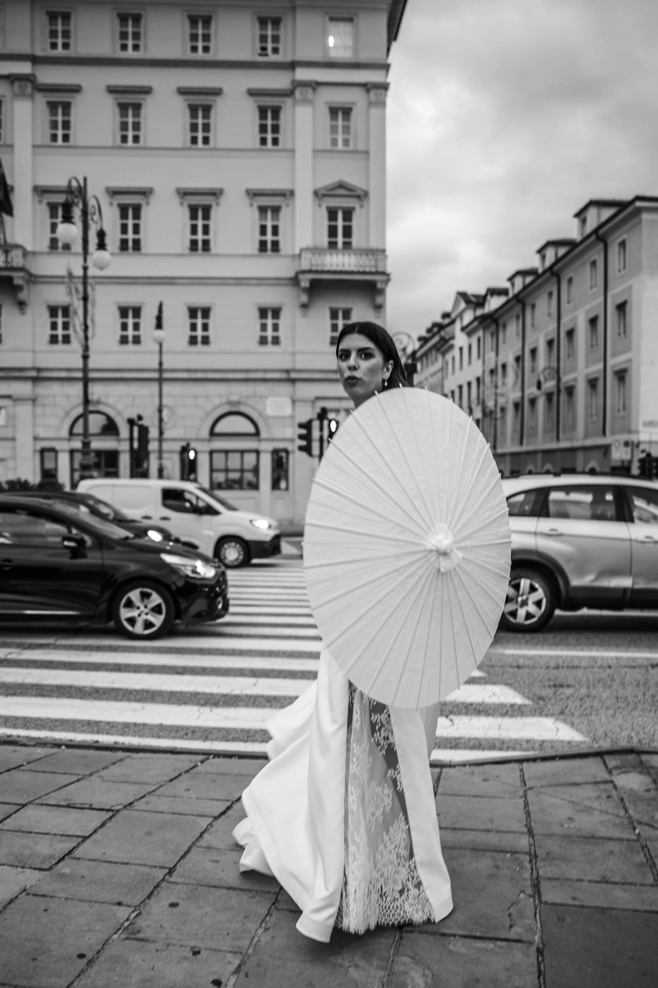 Bride holding a parasol while crossing a street in downtown Trieste during a black and white urban wedding photoshoot in Italy