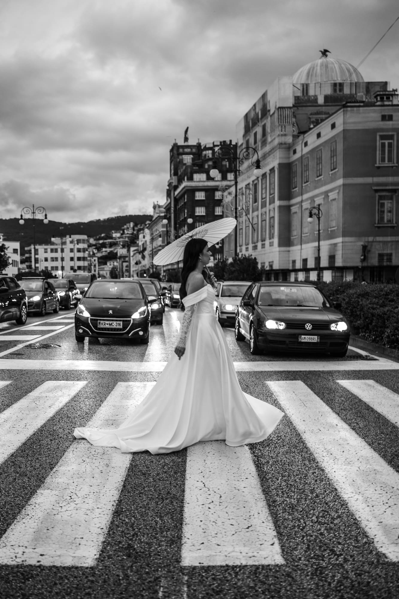 Bride crossing a street in central Trieste with a parasol during a black and white urban wedding photoshoot in Italy