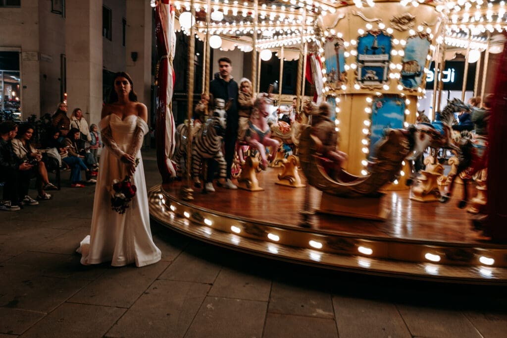 Bride standing beside a vintage carousel in Piazza della Borsa during an evening wedding photoshoot in Trieste, Italy