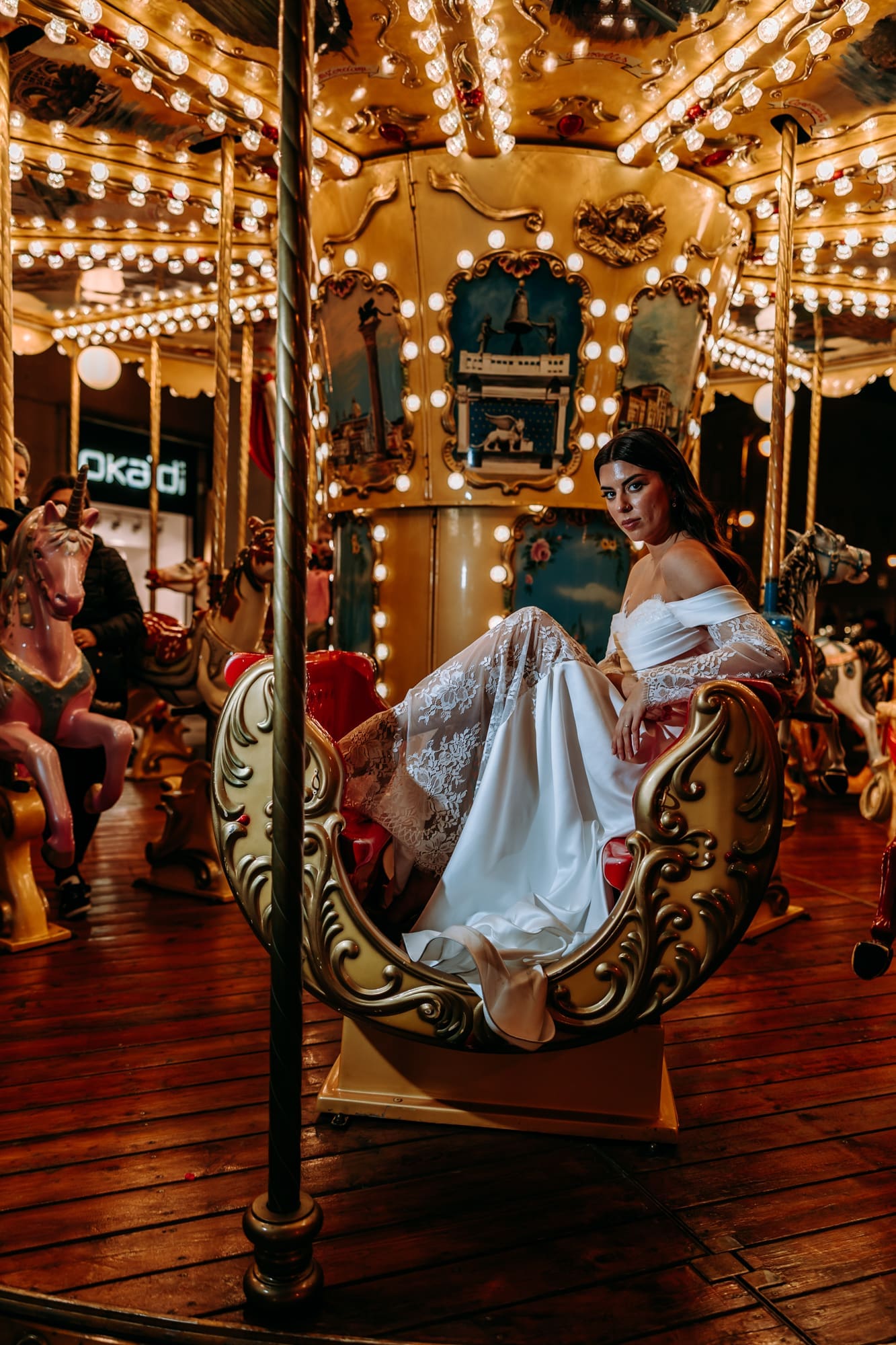 Bride seated in an ornate carousel carriage in Piazza della Borsa during a romantic nighttime wedding photoshoot in Trieste, Italy