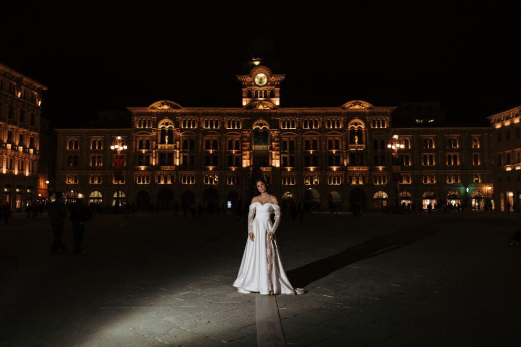 Bride standing in Piazza Unità d’Italia after dark with the illuminated Palazzo del Municipio in the background, Trieste, Italy