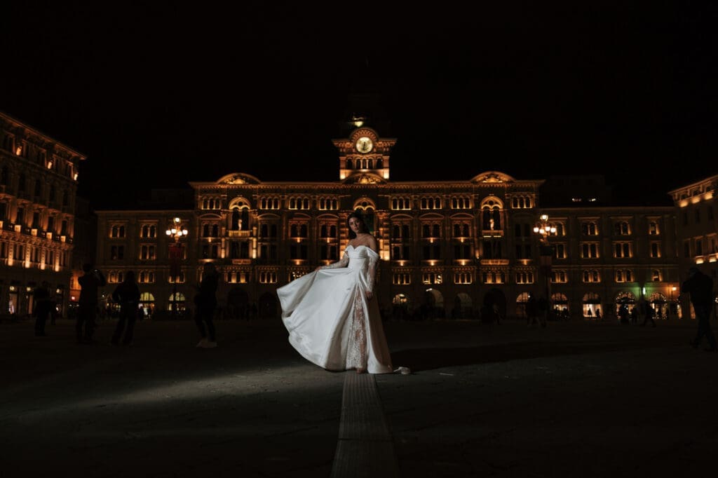 Bride posing in Piazza Unità d’Italia at night with the illuminated Palazzo del Municipio creating a dramatic wedding portrait in Trieste, Italy