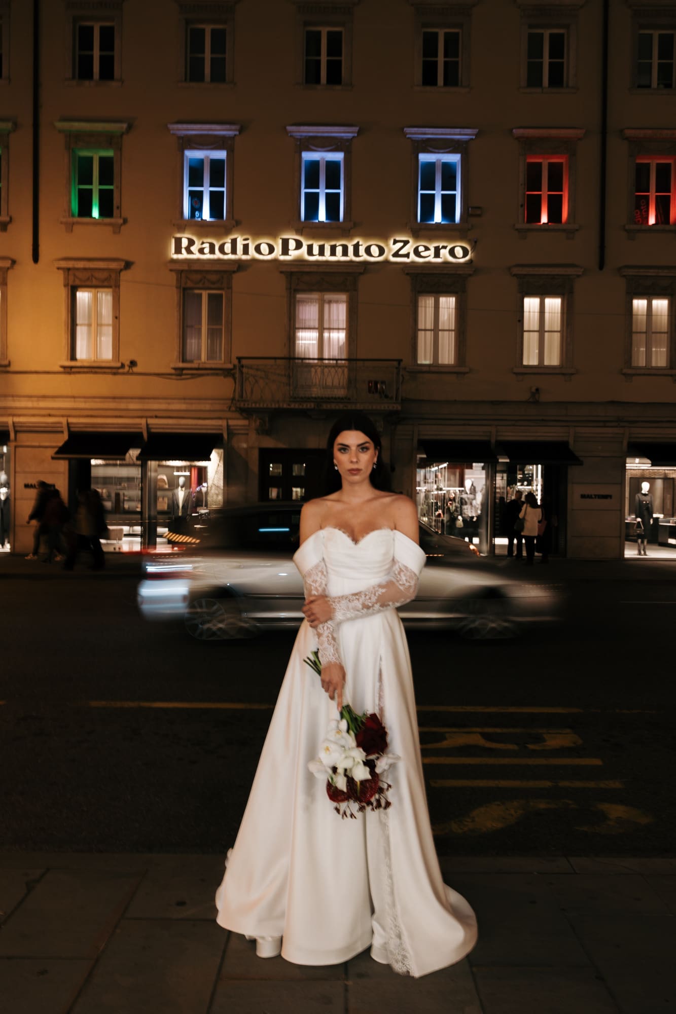 Bride posing in downtown Trieste at night with city lights and motion blur creating a modern editorial wedding portrait in Italy