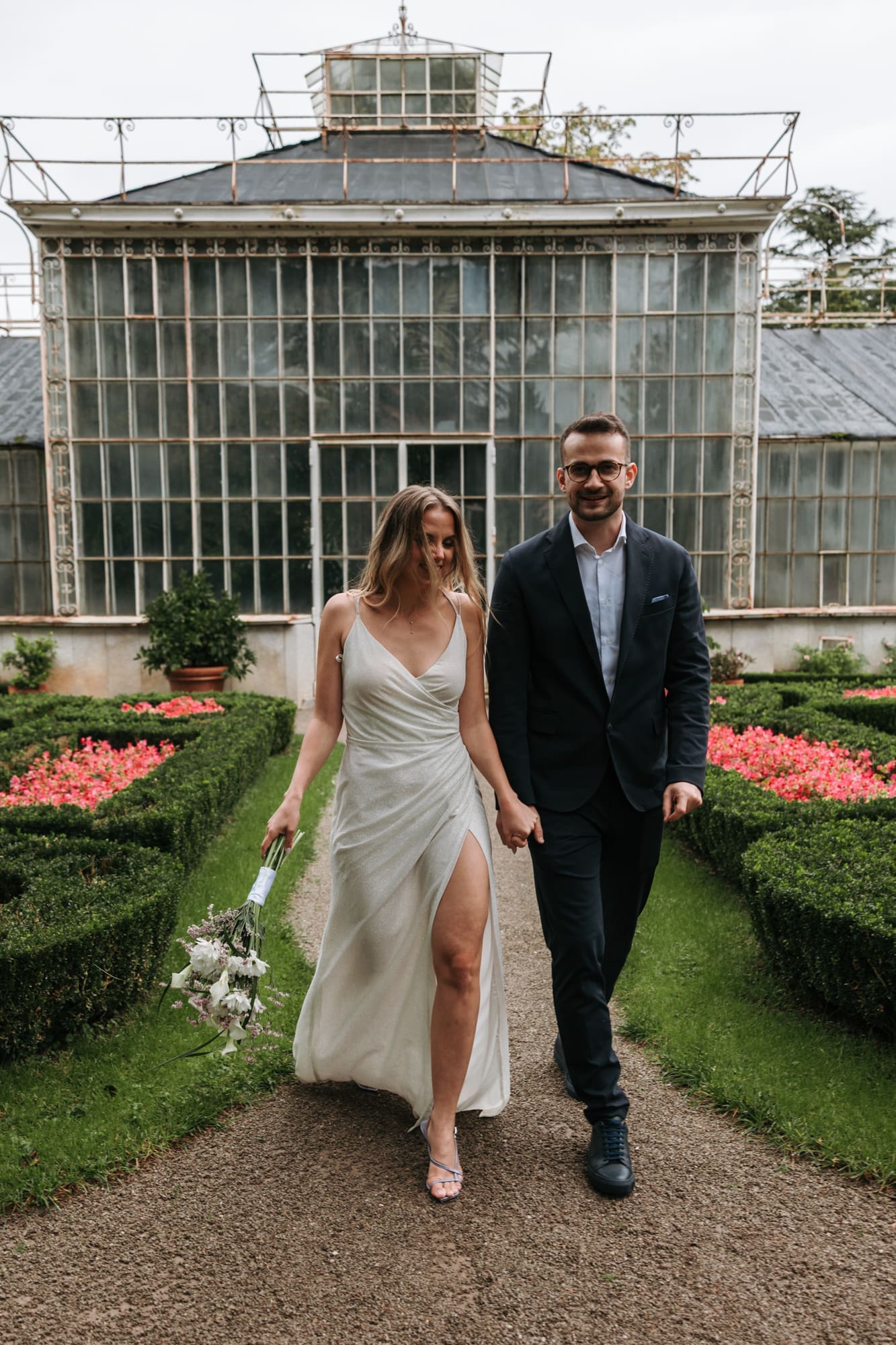 Bride and groom walking through the formal gardens of Villa Mirasasso with the historic greenhouse in the background near Sežana, Slovenia.