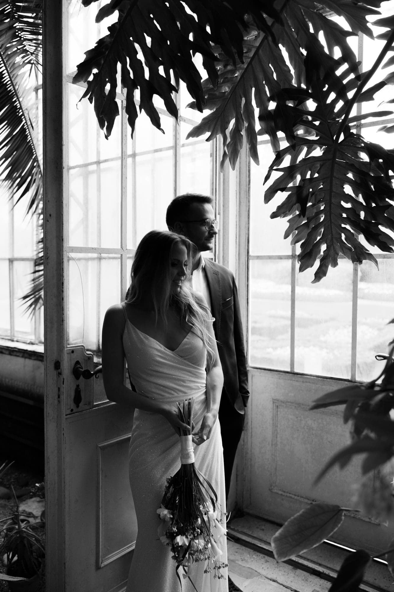 Bride and groom standing by the greenhouse doors surrounded by tropical plants at Villa Mirasasso in Slovenia.
