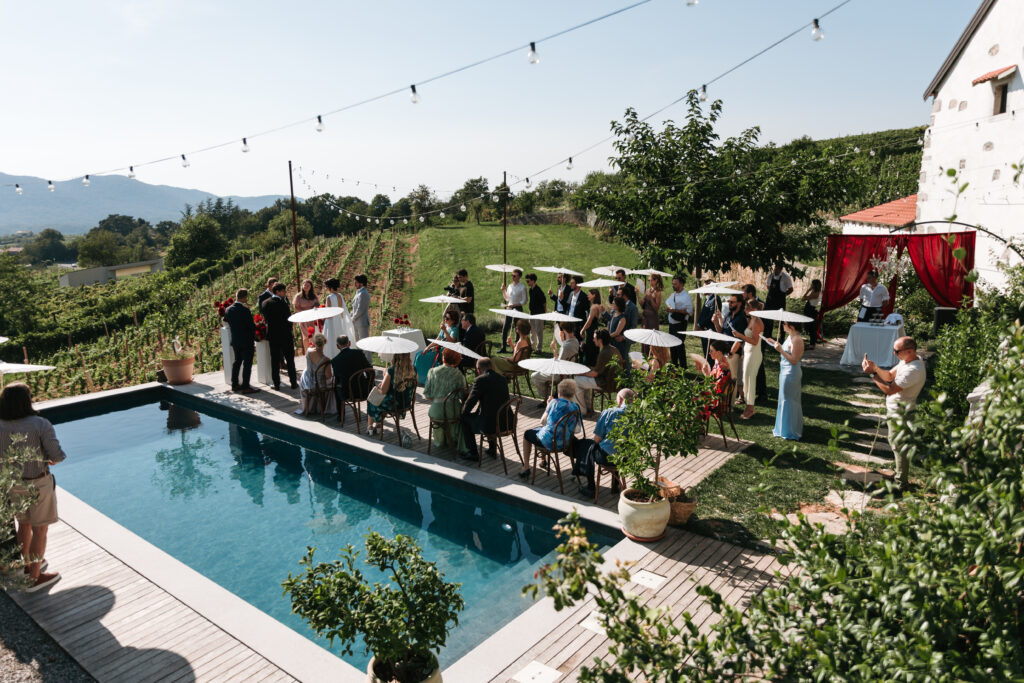 Outdoor wedding ceremony beside a swimming pool, with guests holding white parasols and vineyards stretching across the surrounding hills.