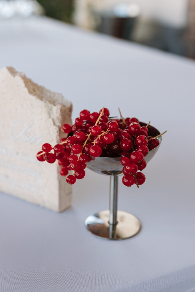 Red currants arranged in a silver pedestal bowl beside a stone sign on a white table at Ajda and Nac’s wedding.