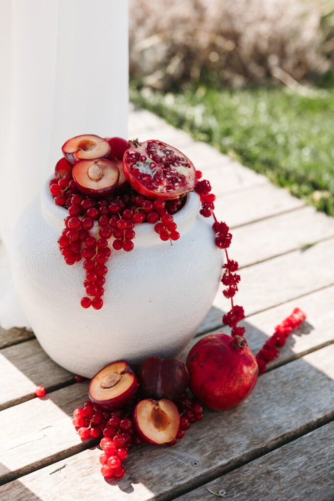 White ceramic vase topped with red currants, halved plums and pomegranate, with more fruit arranged on the wooden deck at Winery Škerlj.