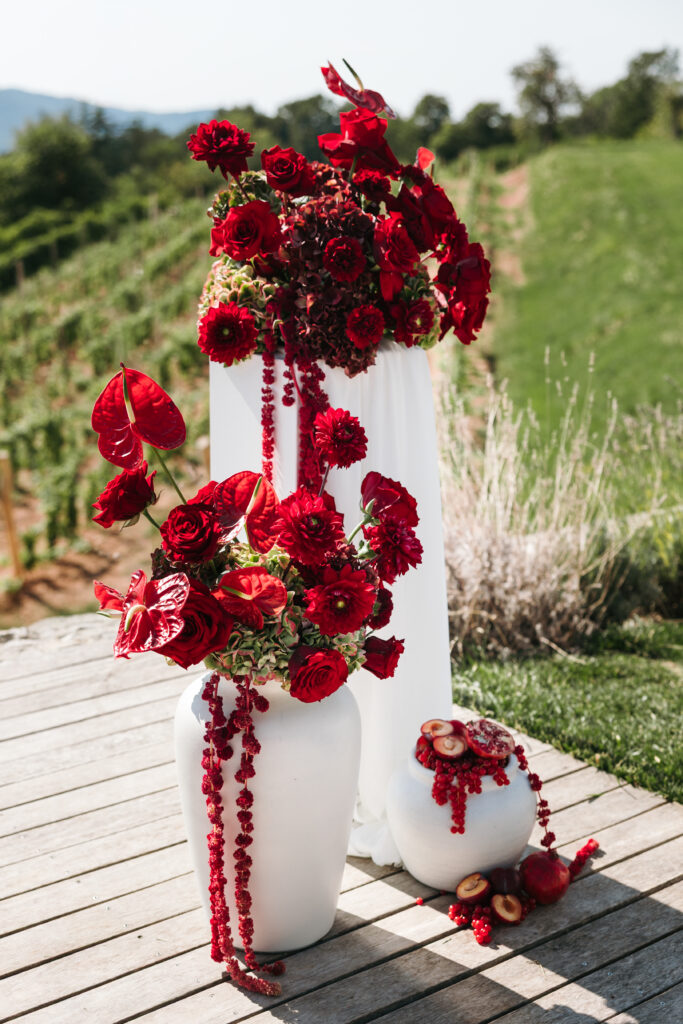 Wedding altar decorated with red flowers and cascading amaranthus in white vases, with fruit accents and vineyards behind at Winery Škerlj.
