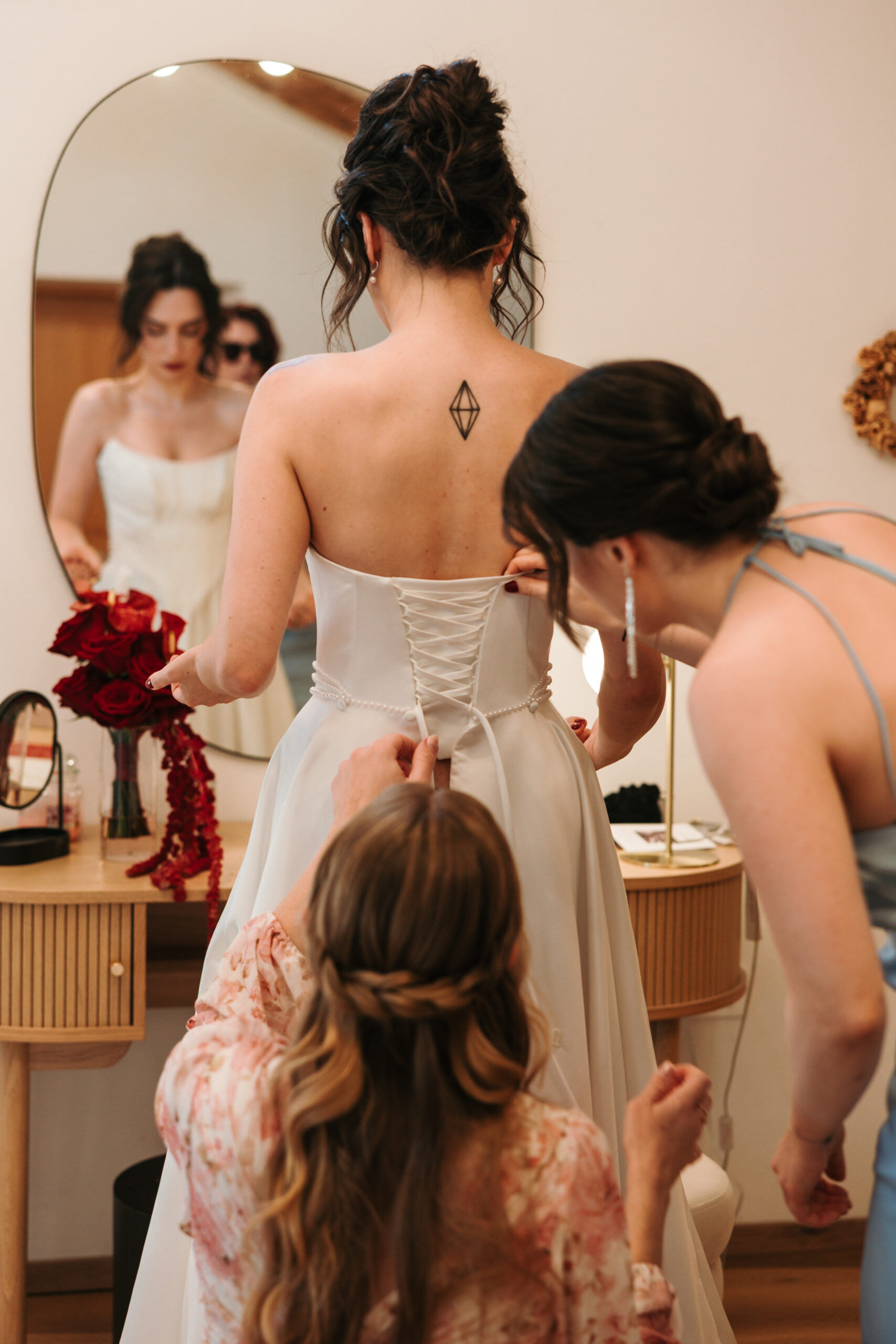 Ajda stands in front of a mirror as two women lace up her white wedding dress during bridal preparations at Winery Škerlj.