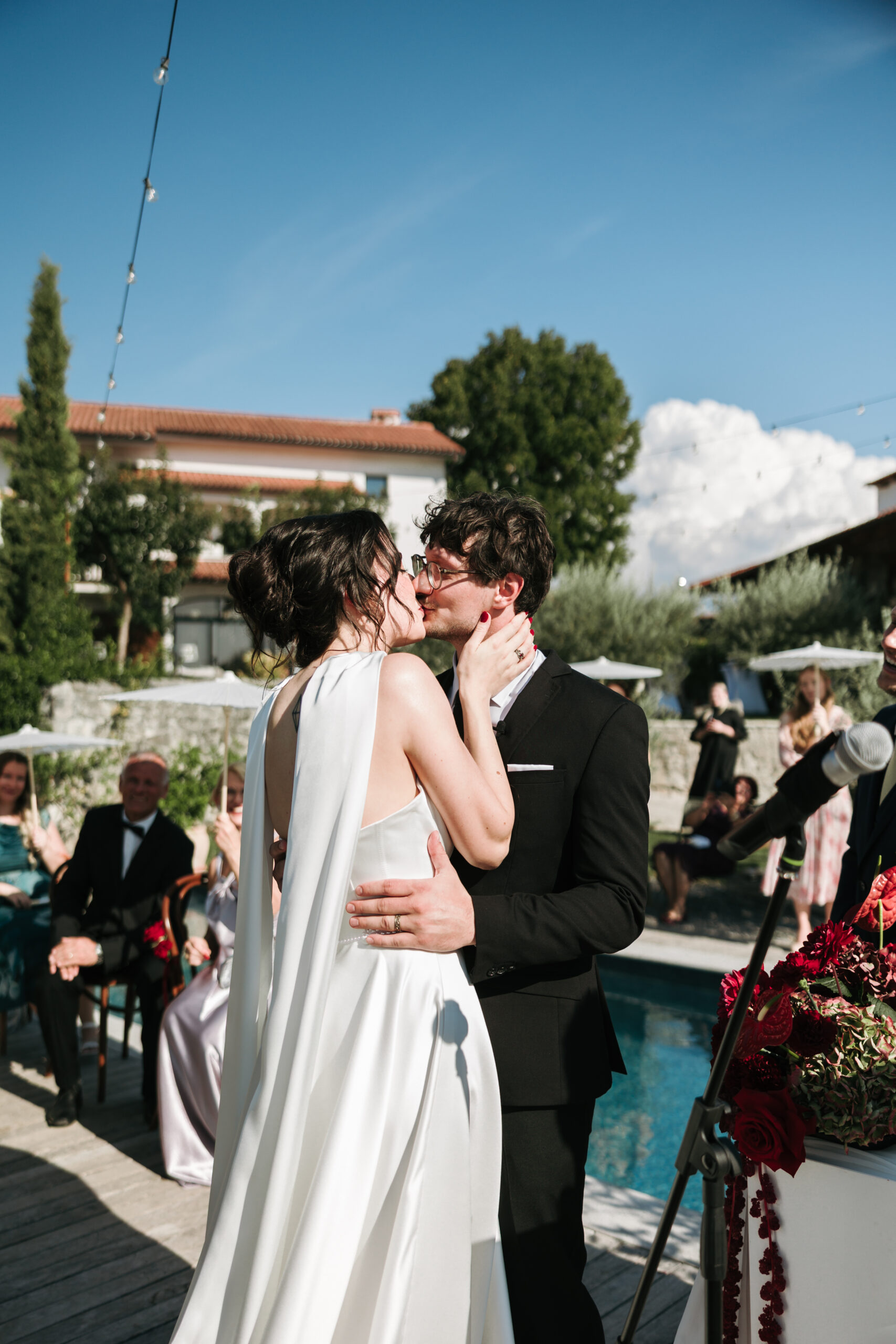 Ajda and Nac kiss during their outdoor wedding ceremony beside the pool at Winery Škerlj, with seated guests and red flowers around them.