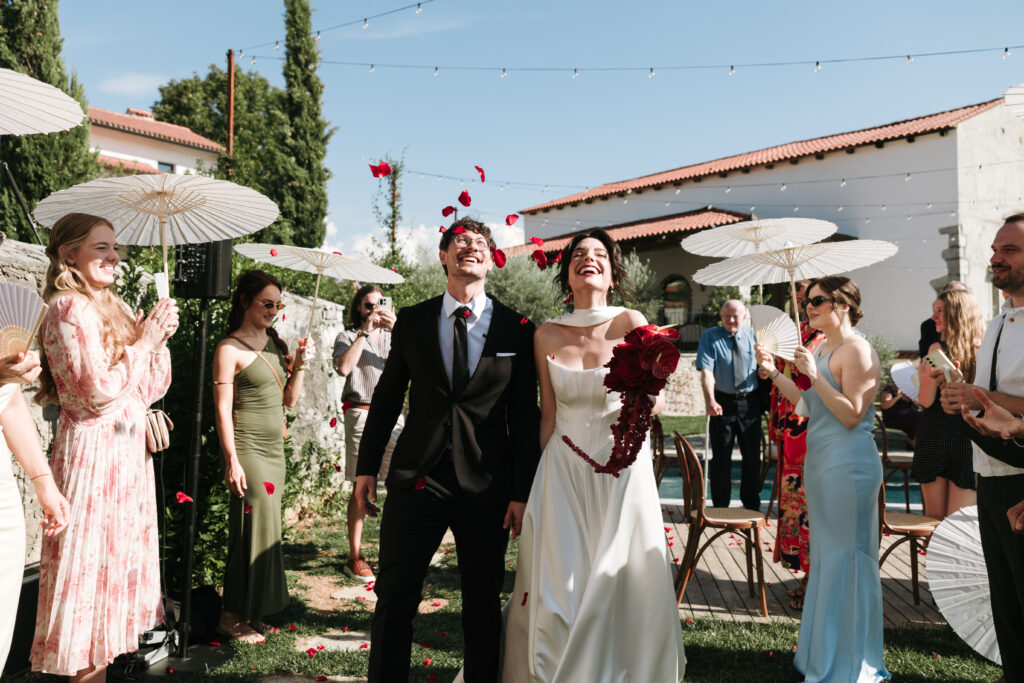 Newlyweds walking hand in hand between applauding guests holding white parasols, with the bride carrying a red bouquet.