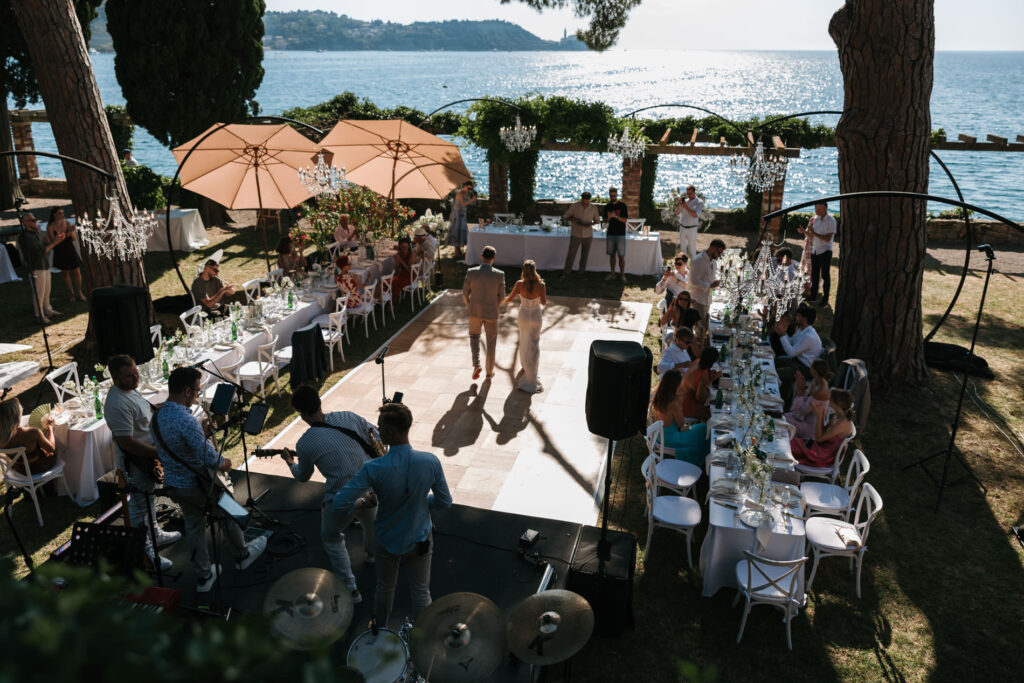 Špela and David walking between long outdoor reception tables at Villa Tartini, surrounded by wedding guests, with live musicians and the sea in the background.