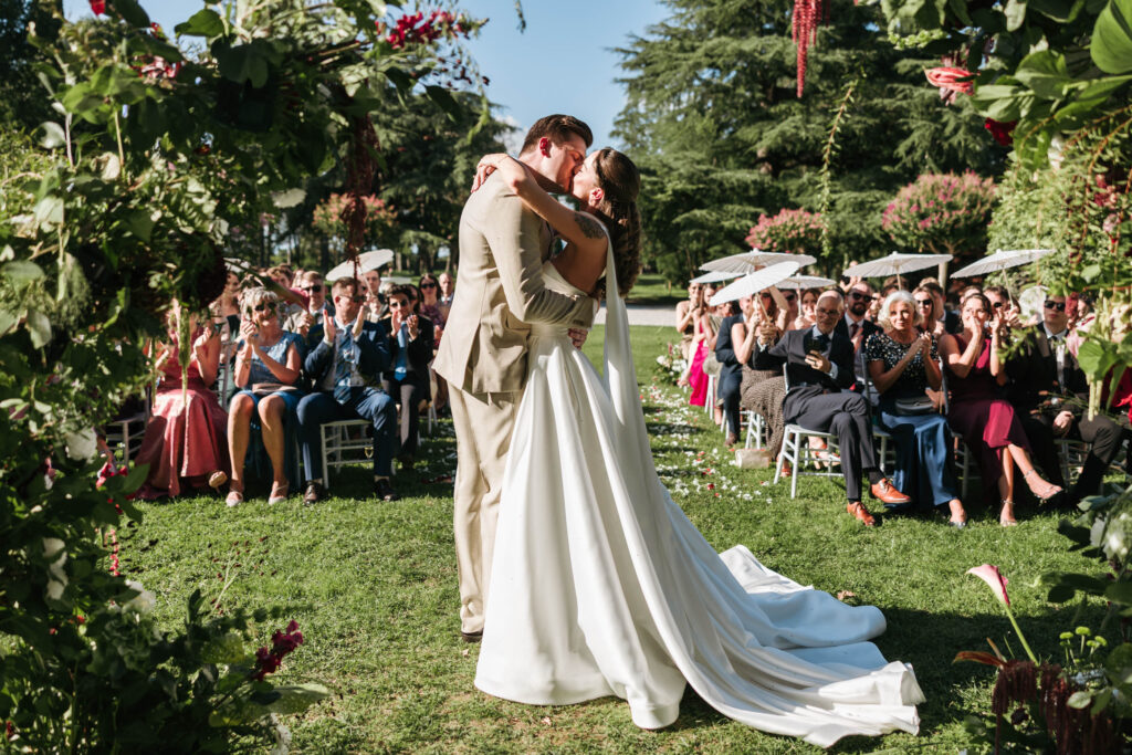 Bride and groom kissing beneath a floral arch during their outdoor wedding ceremony at Villa Locatelli in Italy, with seated guests applauding in the sunlit garden.