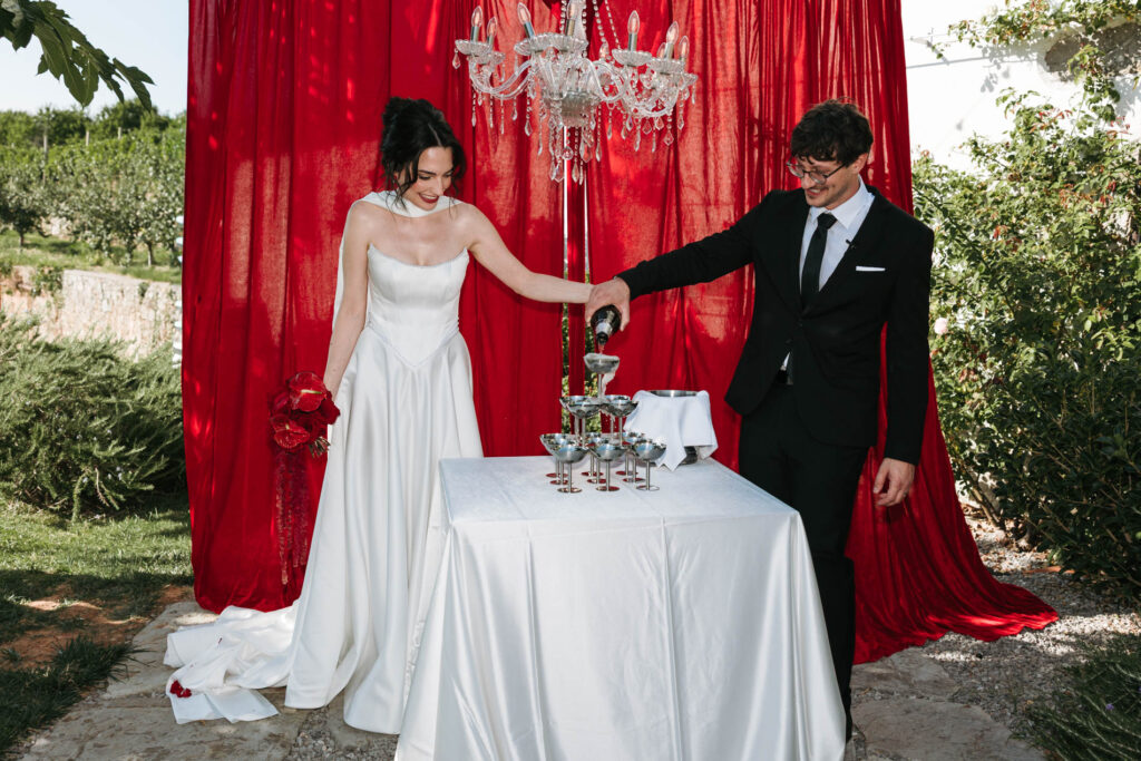 Bride and groom pouring sparkling wine together into a champagne tower, framed by red drapes and a hanging chandelier.