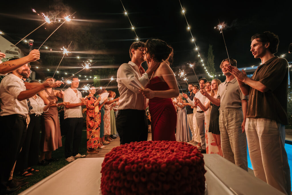 Ajda and Nac kiss beside their red wedding cake as guests hold sparklers under string lights at Winery Škerlj in Slovenia.