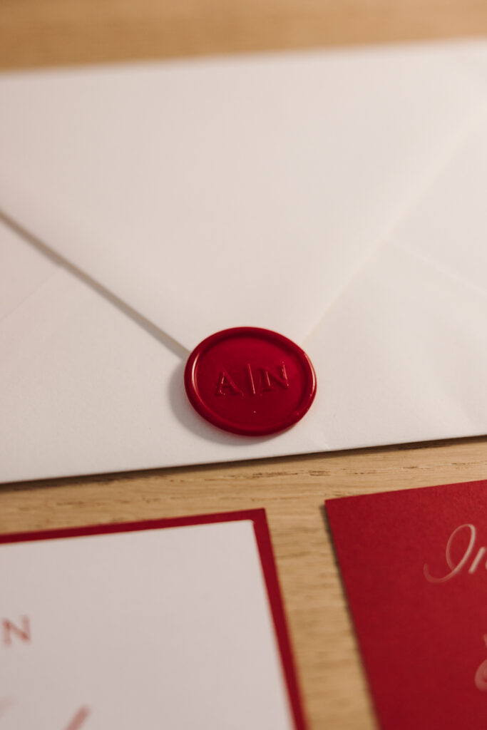 White wedding envelope with a red wax seal bearing Ajda and Nac’s initials, beside red and white stationery.