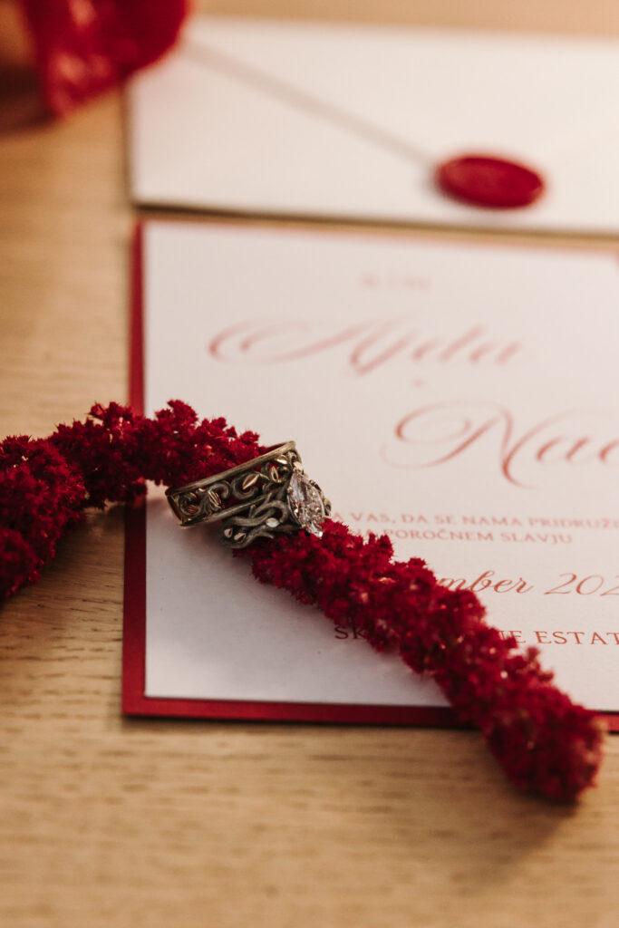 Wedding rings resting on deep red flowers over Ajda and Nac’s red-bordered wedding invitation.