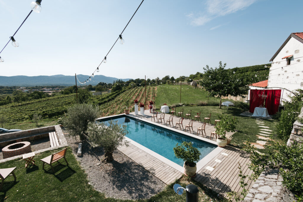 Poolside wedding ceremony setup at Winery Škerlj, with rows of chairs, a red backdrop and views of the surrounding Karst vineyards.