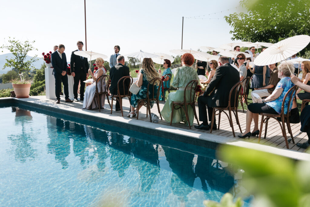 Wedding guests seated beside a swimming pool under white parasols during a sunny outdoor ceremony, with the groom standing at the front.