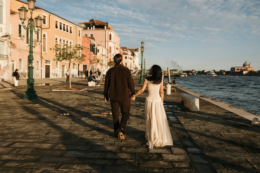 Couple walking hand in hand along the Zattere waterfront in Venice, seen from behind with pastel buildings, ornate lampposts and the Giudecca Canal in afternoon sunlight.